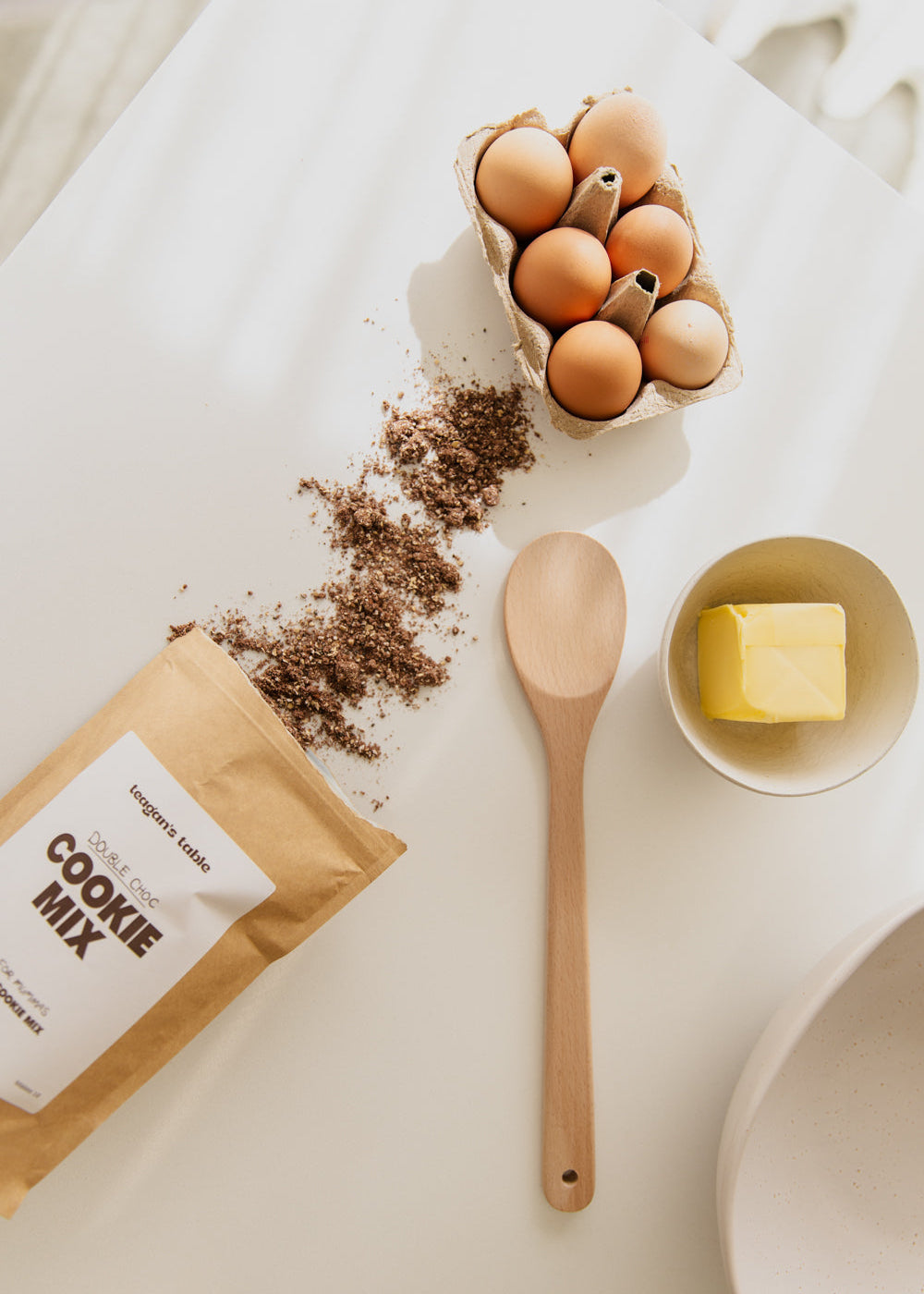 Teagan's Table cookie mix on kitchen bench with eggs and butter and wooden spoon